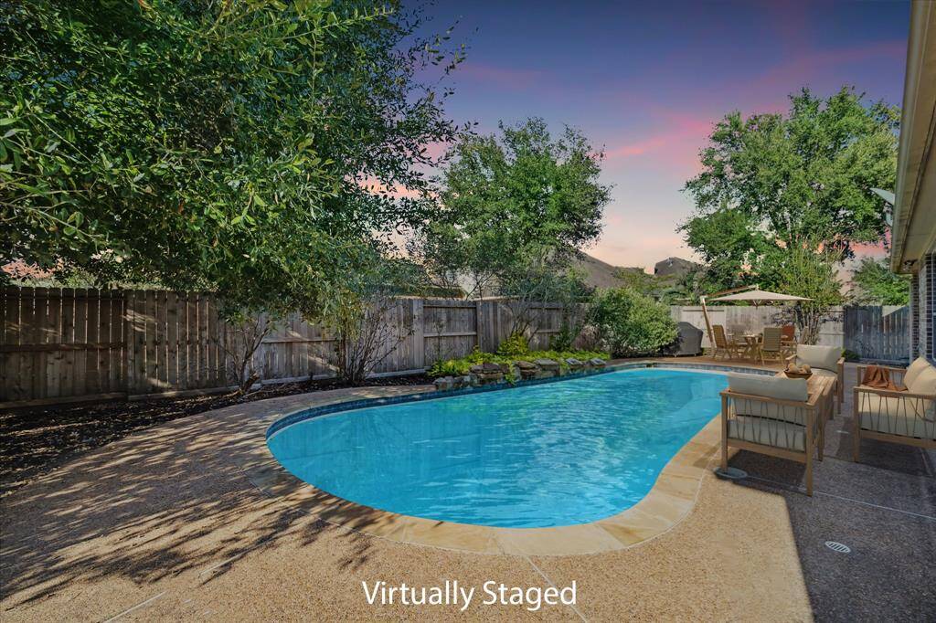 This photo showcases a serene backyard with a large, inviting pool, surrounded by lush greenery and a privacy fence. The space is virtually staged with comfortable outdoor seating, creating a perfect oasis for relaxation and entertaining.