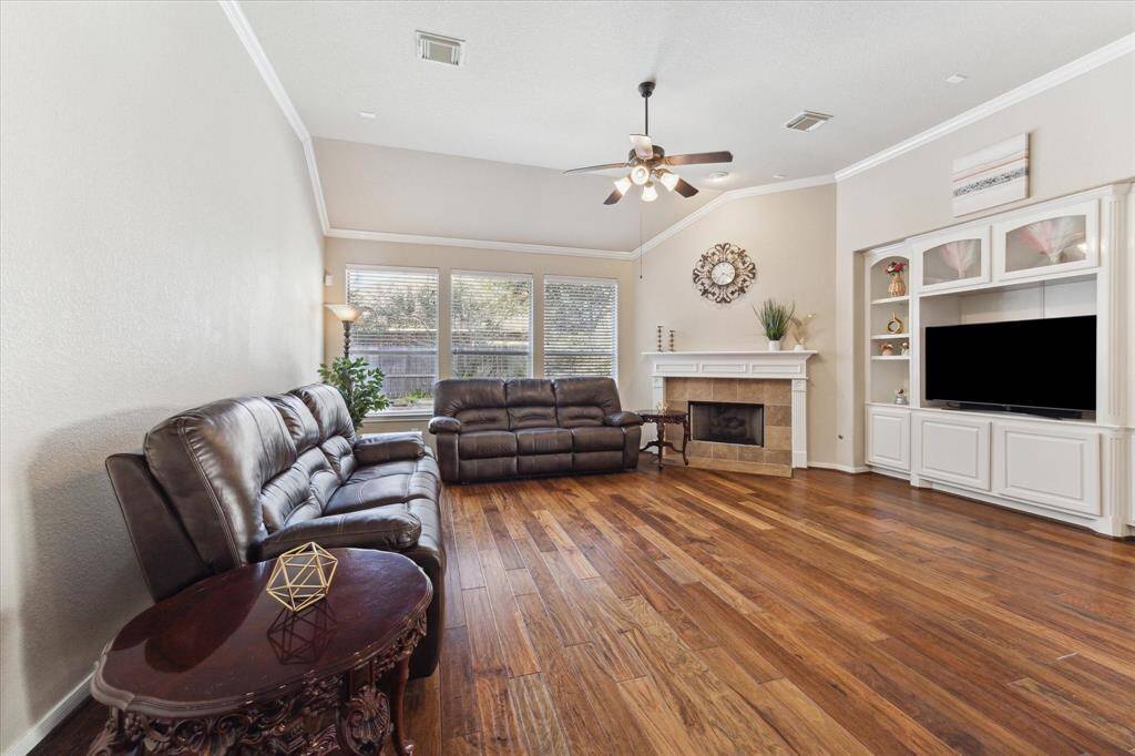 Spacious living room with hardwood floors, high ceiling, and abundant natural light from large windows. Features a cozy fireplace, built-in shelving, and a ceiling fan.