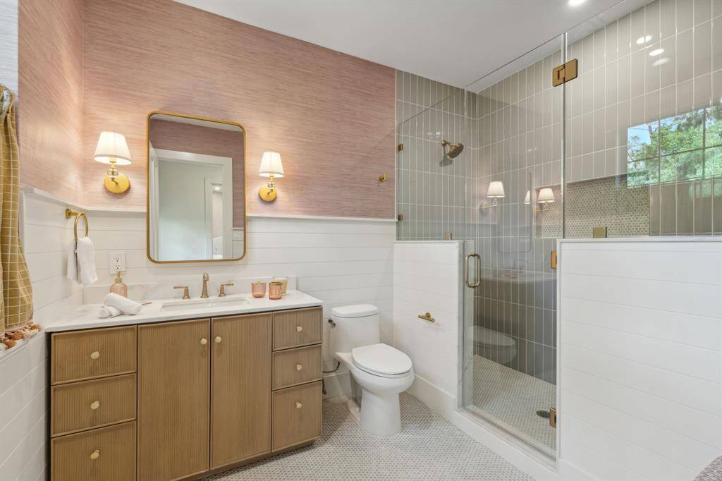 The ensuite bathroom for secondary bedroom #1 offers a ribbed front vanity with electrical outlets & organizers in drawers, complemented by brass plumbing fixtures and a quartz countertop. Decorative sconces, a mirror, and removable pink grasscloth wallpaper create a soft, inviting atmosphere. The walk-in shower w/ a frameless glass wall, bench seat, soap niche with penny tile inlay, and subway tile walls. Not shown is a built-in bench w/custom cushion & new double-pane window w/ a cafe curtain adds natural