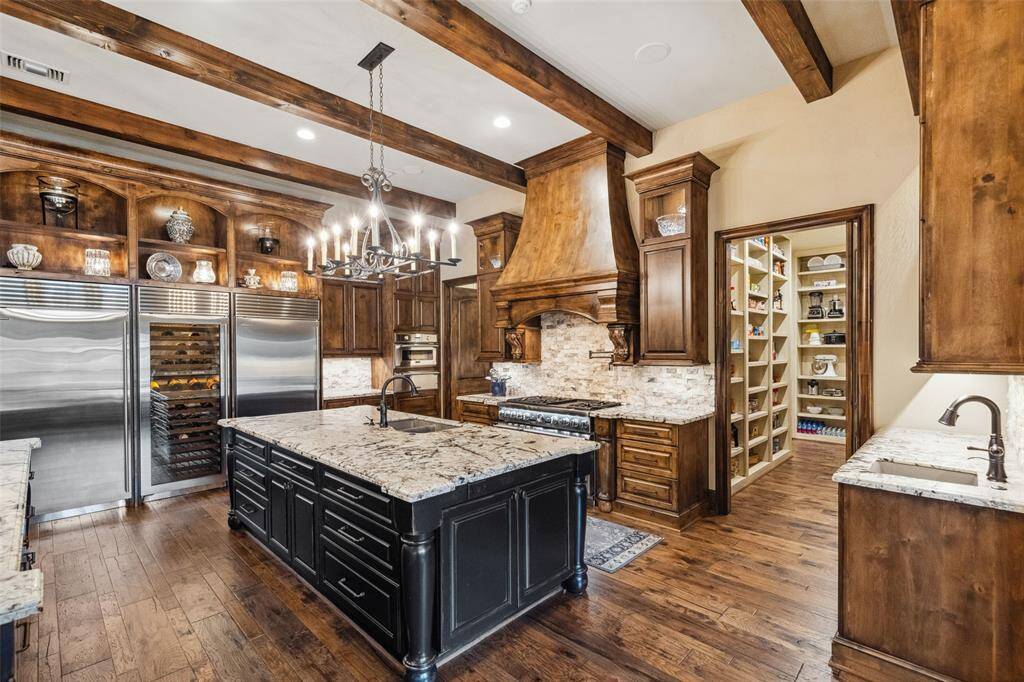 Beautiful iron chandeliers and pendants illuminate the culinary space, drawing attention to the gleaming granite countertops and display items in the open shelving and glass front cabinetry. Repeating timber beams provide additional architectural interest.