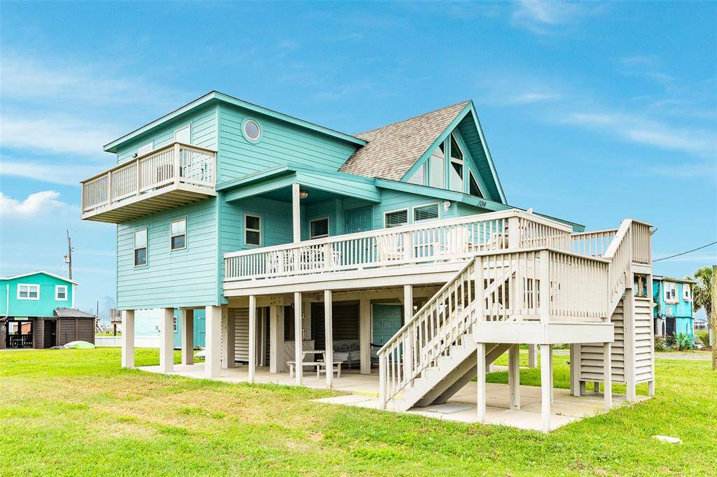 This charming, elevated beach house features a vibrant turquoise exterior with a spacious wraparound deck, perfect for enjoying coastal views. The home includes multiple windows for natural light and a second-story balcony for added outdoor space. Ideal for beachside living.