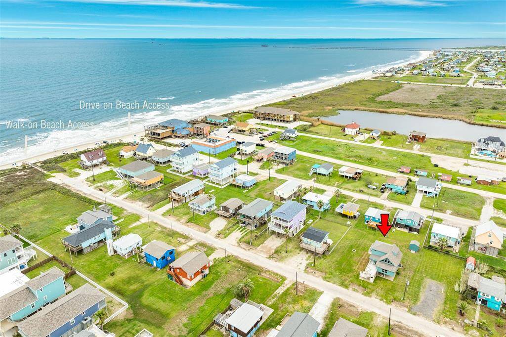This aerial photo showcases a coastal neighborhood with a house marked by a red arrow. The area features easy access to the beach, with both drive-on and walk-on paths visible nearby. The homes are situated close to the shore, offering potential ocean views and a relaxed seaside lifestyle.