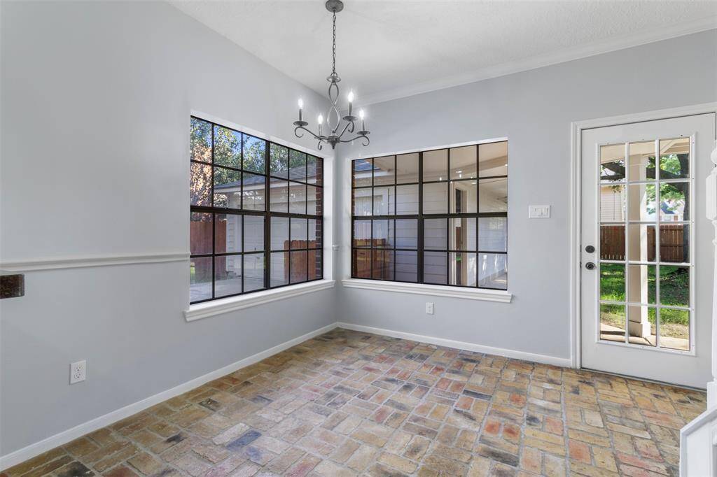 Bright dining nook with large windows, brick flooring, and a stylish chandelier. A glass door leads to a lush backyard, providing ample natural light and charming views.