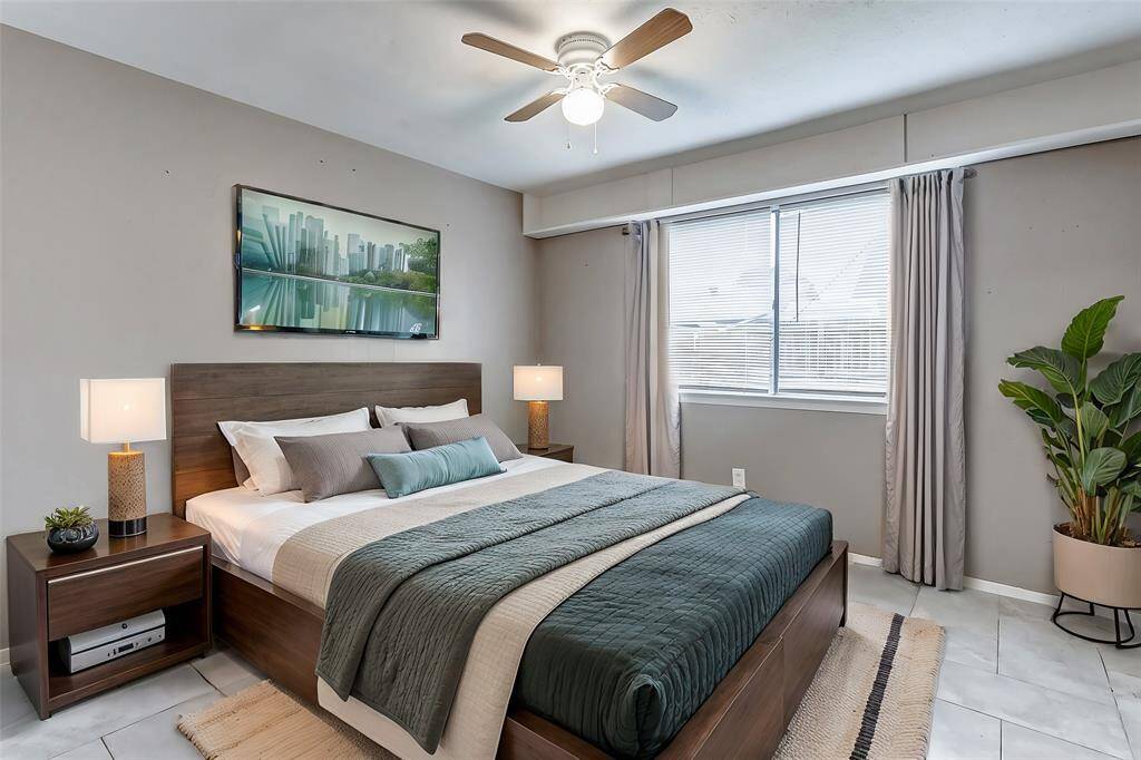 This primary bedroom boasts a serene atmosphere with its neutral palette and modern tile flooring. Natural light flows through the large window, creating a warm and inviting retreat. A ceiling fan enhances comfort, making this space a peaceful escape.