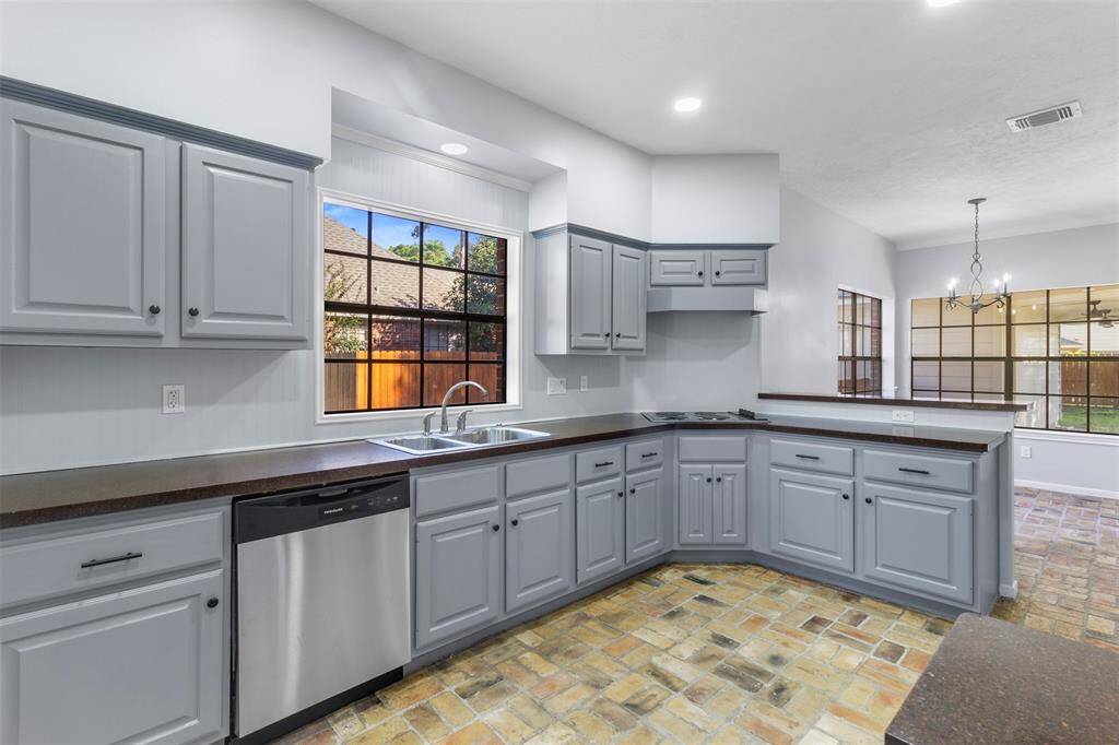 This photo showcases a spacious kitchen with light gray cabinetry, dark countertops, and stainless steel appliances. Large windows provide natural light, and the open layout connects to a dining area with brick flooring.