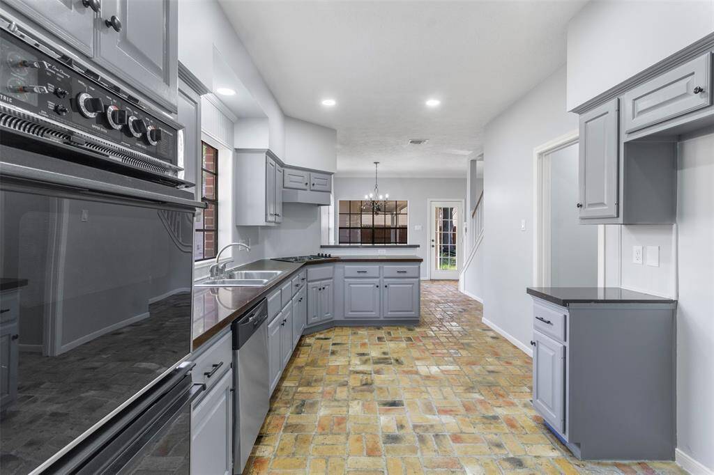 This kitchen features sleek gray cabinetry, modern appliances, and a brick tile floor. It offers ample counter space and opens into a dining area with a view of a glass-paneled door and staircase, creating a bright and inviting atmosphere.