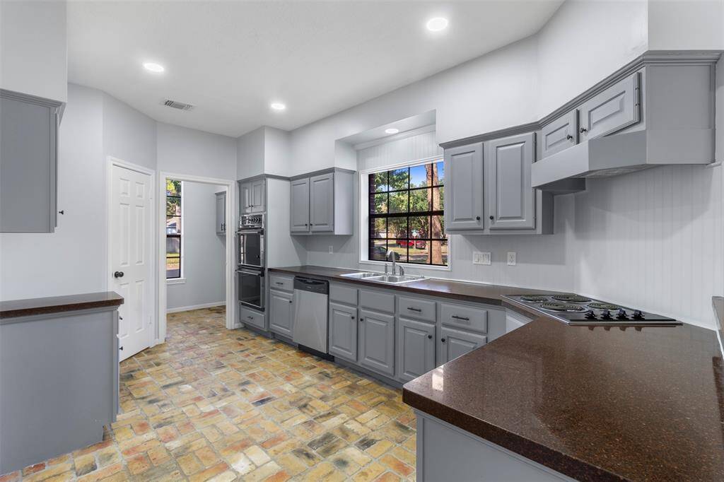 This kitchen features modern gray cabinetry, dark countertops, and a brick-style floor. It includes a built-in oven, stovetop, and dishwasher. A large window offers natural light and a view of the outdoors.