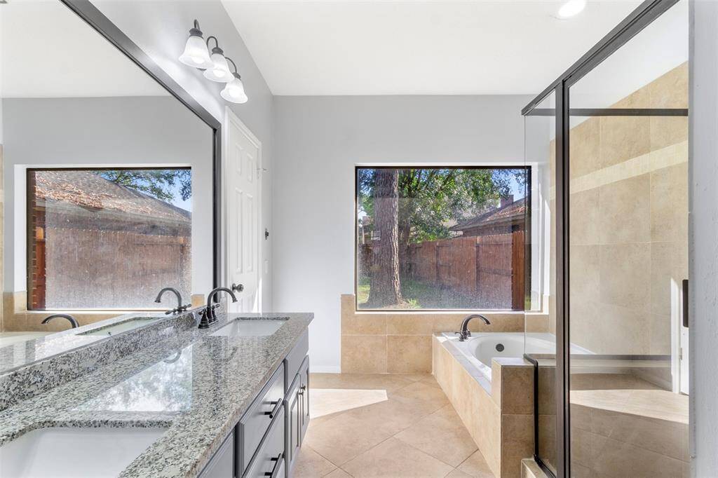 This modern Master bathroom features a spacious layout with a large granite double vanity, a glass-enclosed shower, and a separate soaking tub. Natural light fills the room through two large windows, enhancing the elegant tile design.
