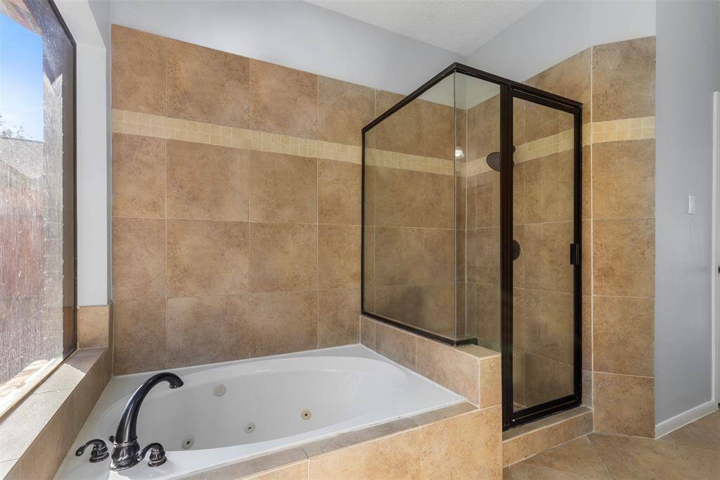 This bathroom features a spacious glass-enclosed shower and a relaxing jetted bathtub, surrounded by elegant beige tiles and natural light from a large window.