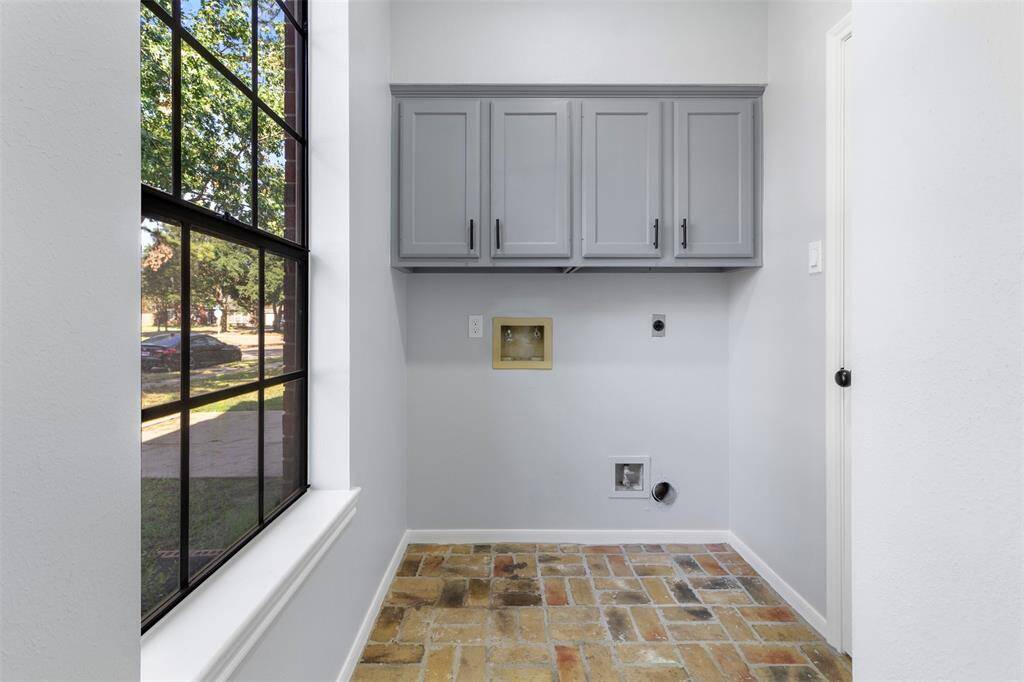 A bright utility room with large windows, brick flooring, and gray cabinets. Ideal for laundry with plumbing and electrical hookups.