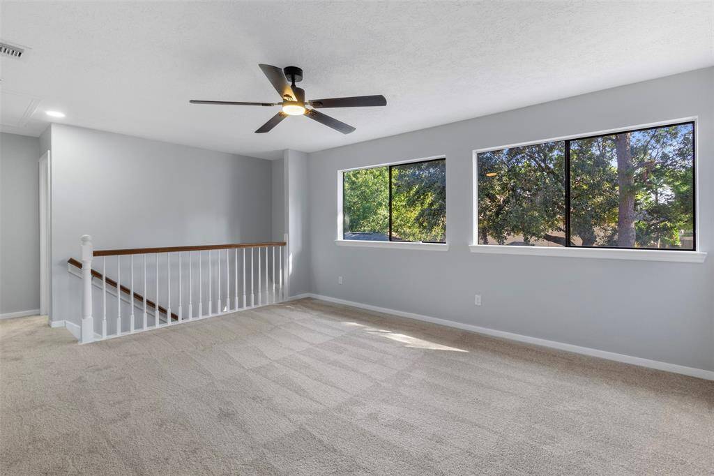 Bright and spacious Game room with neutral carpet, large windows offering lush views, and a modern ceiling fan. An open staircase with a white railing adds a touch of elegance, and the soft gray walls enhance the airy feel.