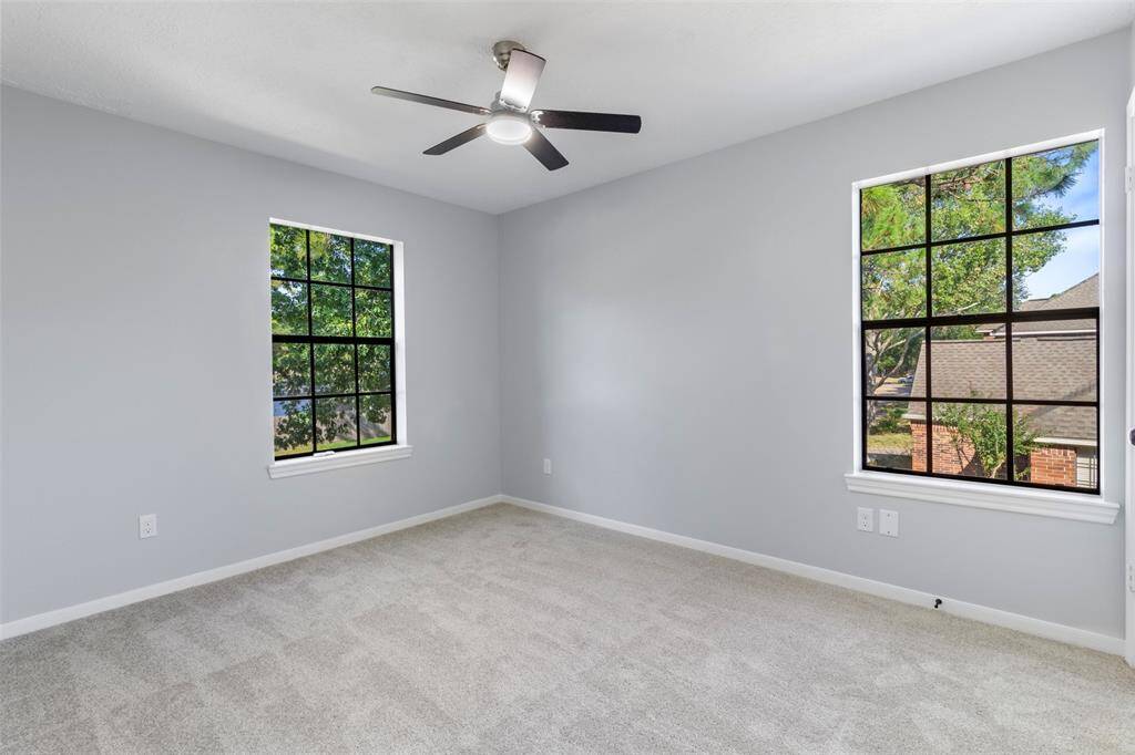 Bright, spacious room with neutral gray walls, plush carpet, and a modern ceiling fan. Two large windows provide ample natural light and a pleasant view.