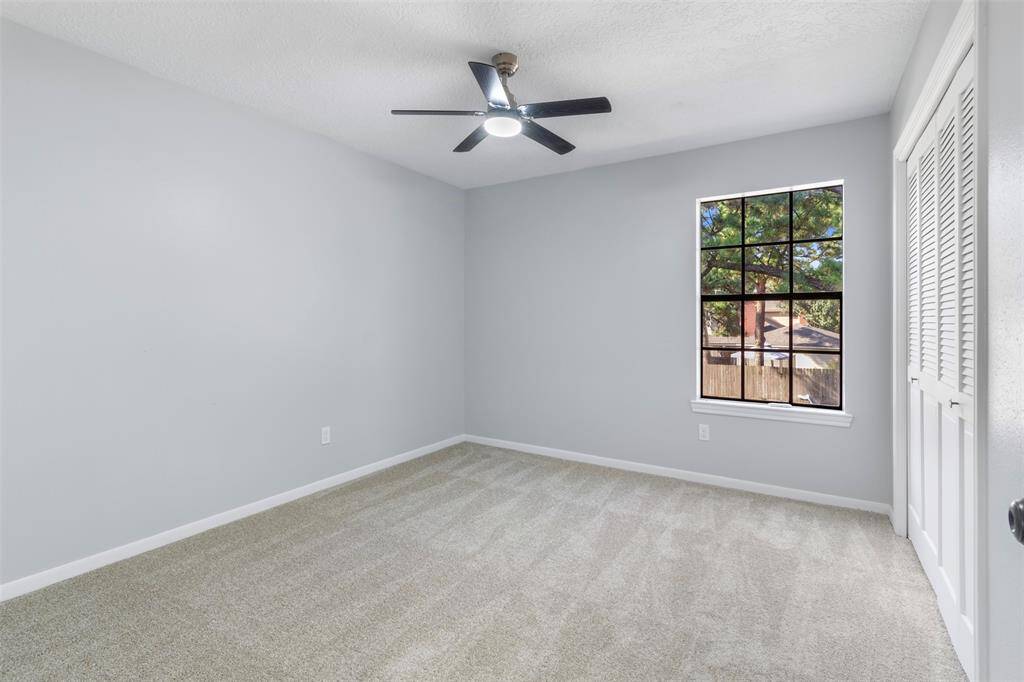 This room features a neutral color palette with light gray walls and carpeted flooring. It includes a ceiling fan and a window offering natural light, as well as a closet with louvered doors.