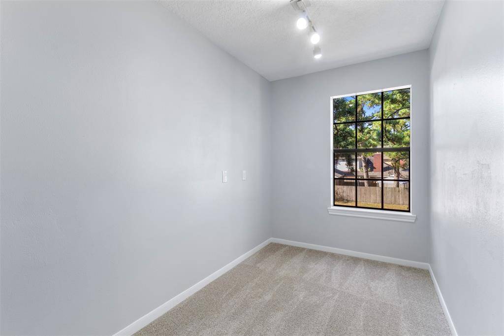 This room features soft gray walls, a large window with a view of greenery, and plush carpeting. It's well-lit with modern ceiling lights, creating a cozy, inviting space suitable for a small office or reading nook.