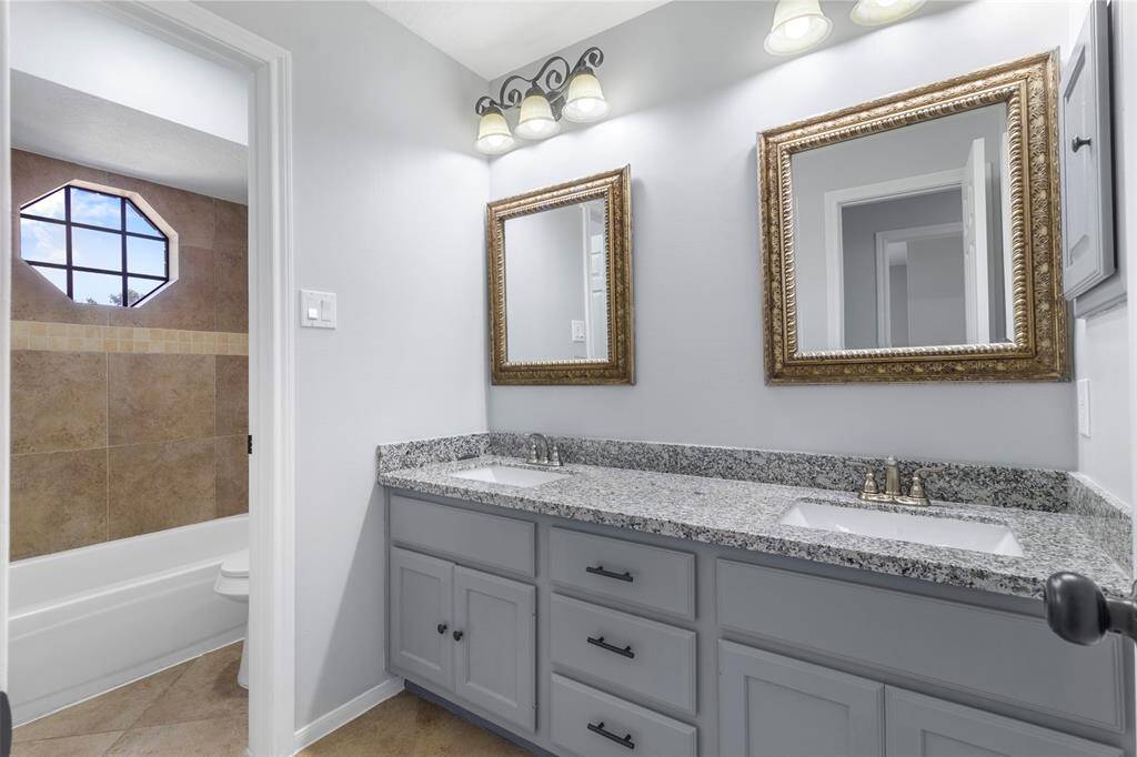 This bathroom features a double vanity with granite countertops, stylish framed mirrors, and modern lighting. Adjacent is a tiled bath area with a unique octagonal window, providing natural light and a touch of elegance.
