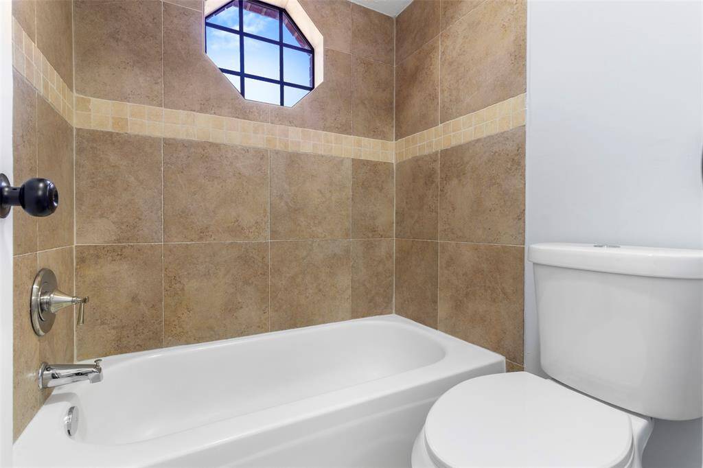 This bathroom features a modern tub-shower combo with beige tiled walls, a small window for natural light, and a clean, white toilet.