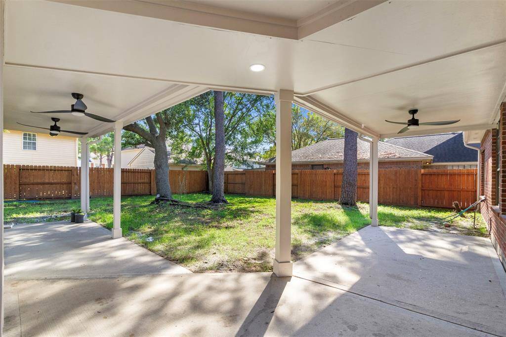 This photo showcases a spacious covered patio with ceiling fans, overlooking a fenced backyard with mature trees, providing a comfortable outdoor living space.