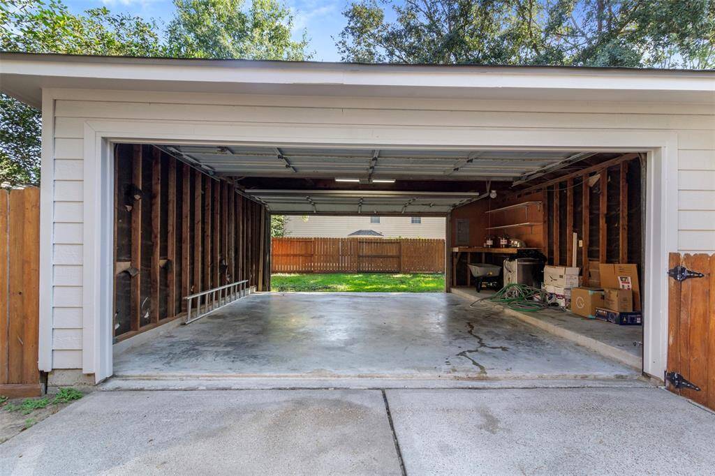 This photo shows a spacious two-car garage with an open door, providing a view of a fenced backyard. The garage has exposed wooden beams, shelving, and space for storage or a workshop.