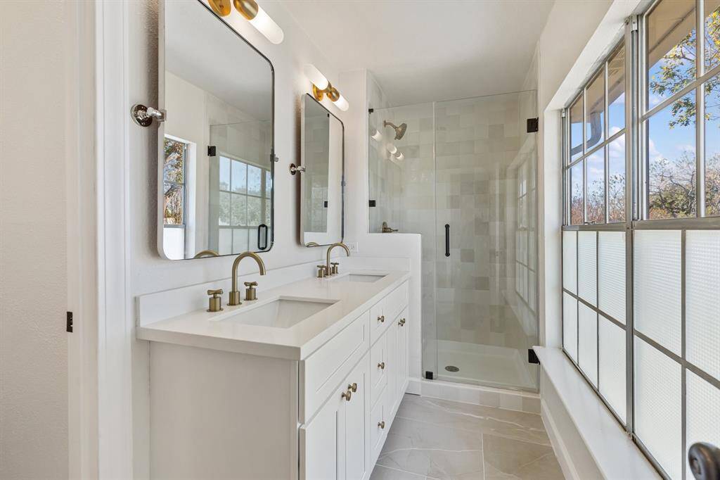 Primary bathroom features a modern design with a double vanity, sleek gold fixtures, large mirrors, and a spacious glass-enclosed shower. Ample natural light, enhancing the bright and airy atmosphere.