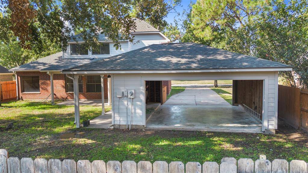 This photo shows a spacious backyard with a two-car carport and a partial view of a two-story brick house. The area is surrounded by trees, providing a sense of privacy and shade.