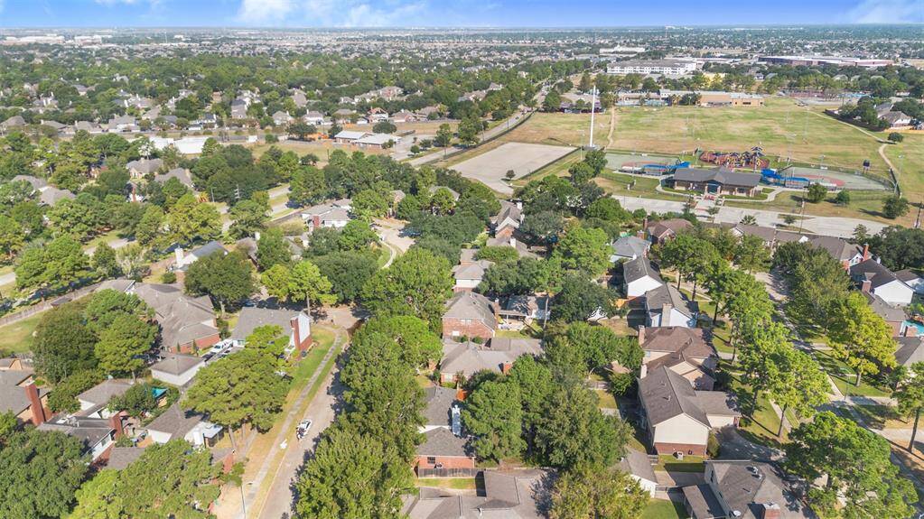 This photo shows a suburban neighborhood with tree-lined streets and well-spaced homes. In the background, there's a large park with a playground and sports facilities, providing ample recreational opportunities. The area is lush and green, offering a peaceful residential setting.