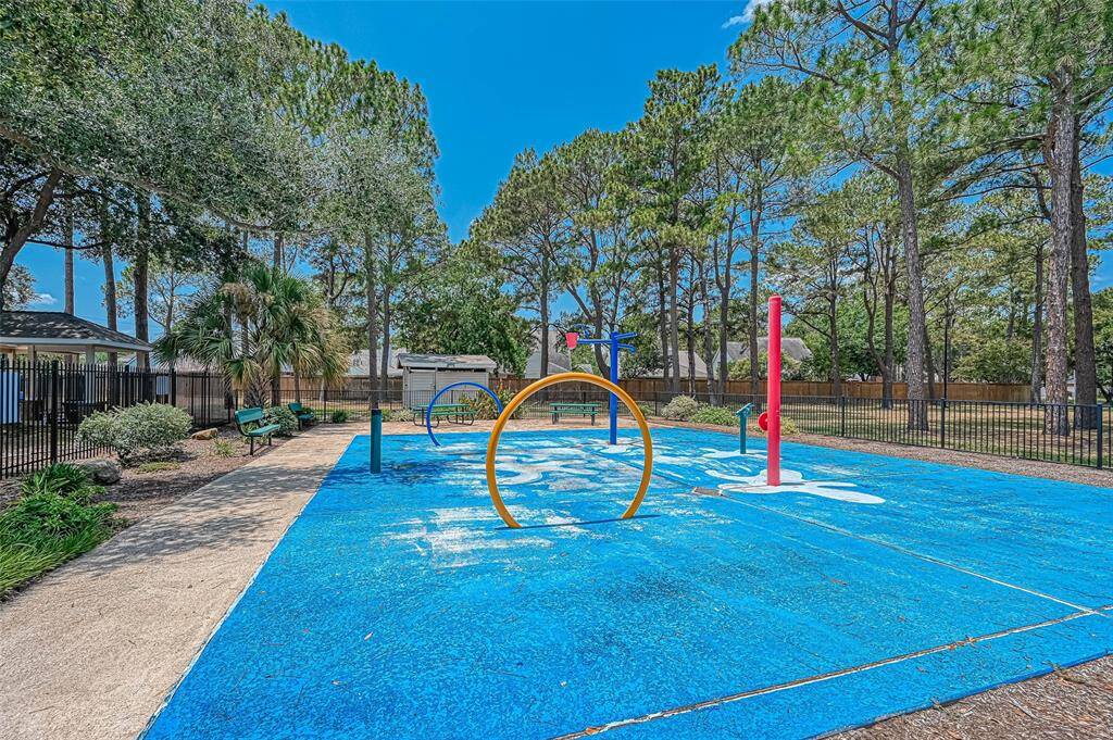 This photo features a vibrant splash pad in a community area, surrounded by trees and a fenced perimeter. Ideal for families, it offers a fun outdoor space for children to play.