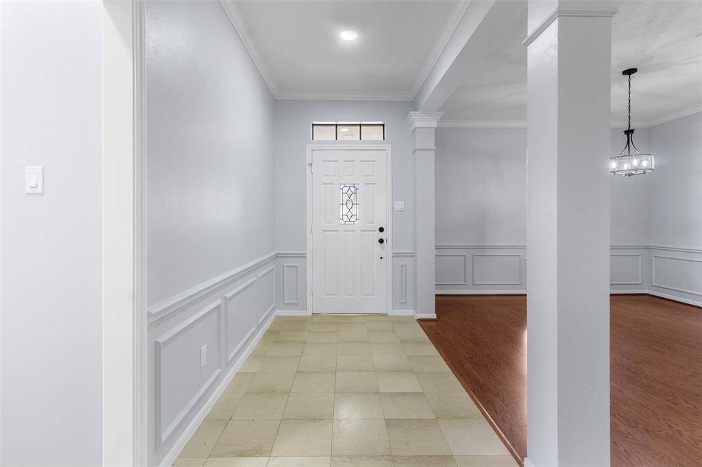 Bright entryway with a white door, decorative glass, and elegant wainscoting. The area features tiled flooring leading to a spacious room with hardwood floors and a modern chandelier.