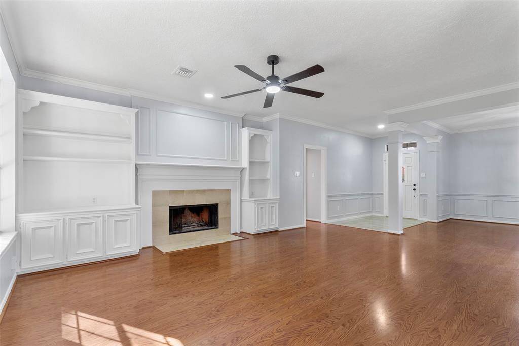 Bright living room with hardwood floors, a cozy fireplace, built-in shelves, and ceiling fan. Features elegant wainscoting and ample natural light. Perfect for entertaining or relaxing.