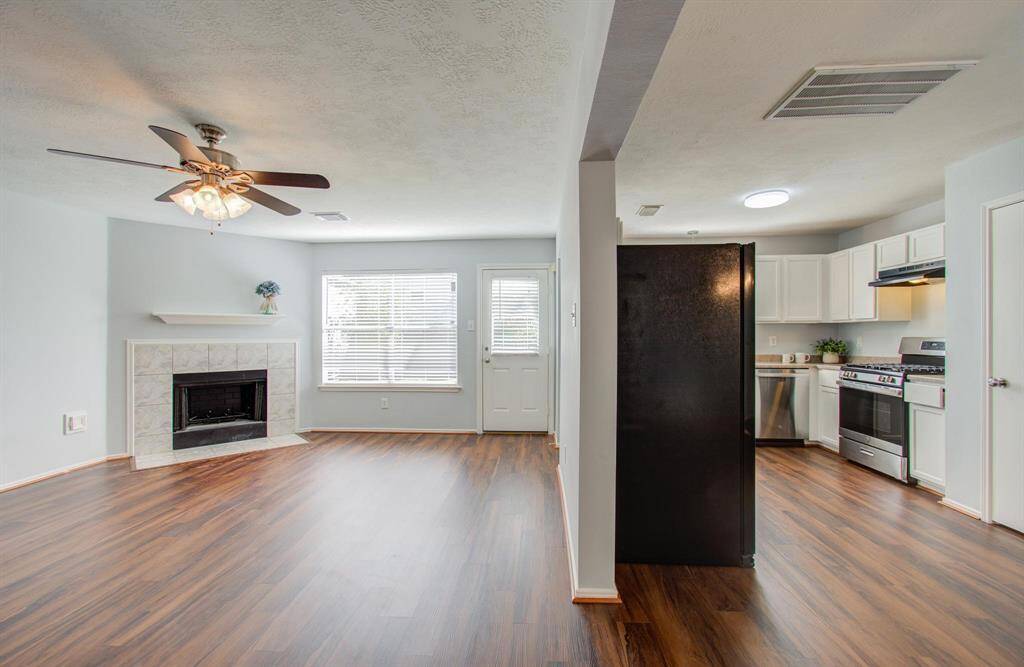 This photo shows a bright, open-concept space with vinyl floors, featuring a cozy living area with a fireplace, and an adjacent modern kitchen with white cabinets and stainless-steel appliances.