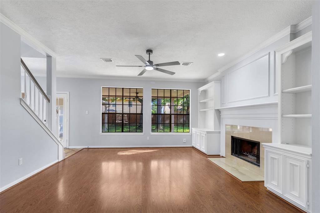 This spacious living room features elegant wood flooring, a cozy fireplace, and built-in shelves. Large windows provide ample natural light, while a ceiling fan offers additional comfort. The room has a fresh, modern look with light-colored walls and trim.