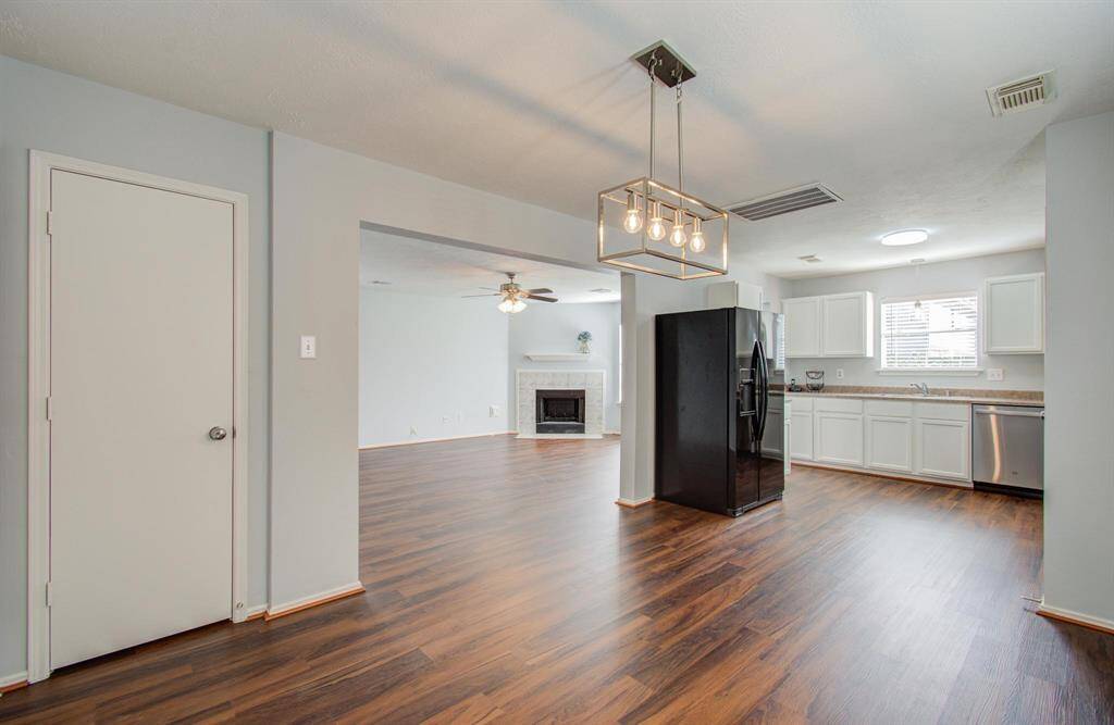 This photo showcases an open-concept living area with a modern kitchen featuring white cabinetry and stainless-steel appliances. The space has vinyl flooring, a cozy fireplace, and a stylish light fixture, offering a bright and inviting atmosphere.
