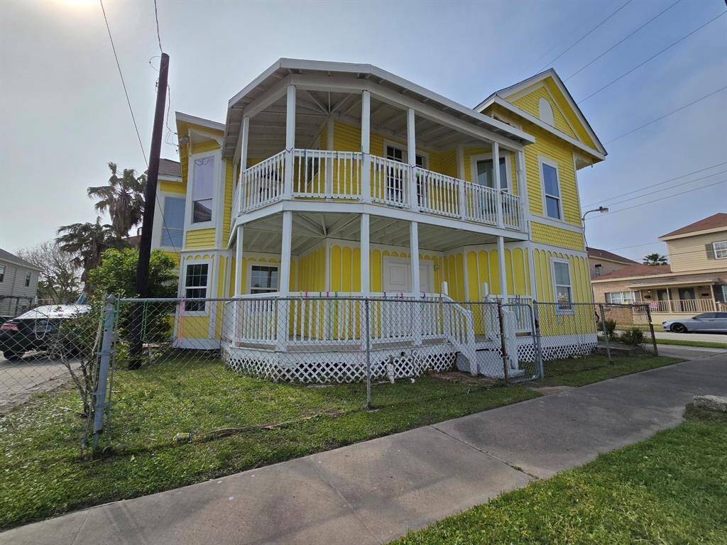This charming, two-story yellow home features a wrap-around porch, ample windows for natural light, and a fenced yard. It has a driveway for parking and is situated on a corner lot with palm trees, offering a vibrant and inviting exterior.