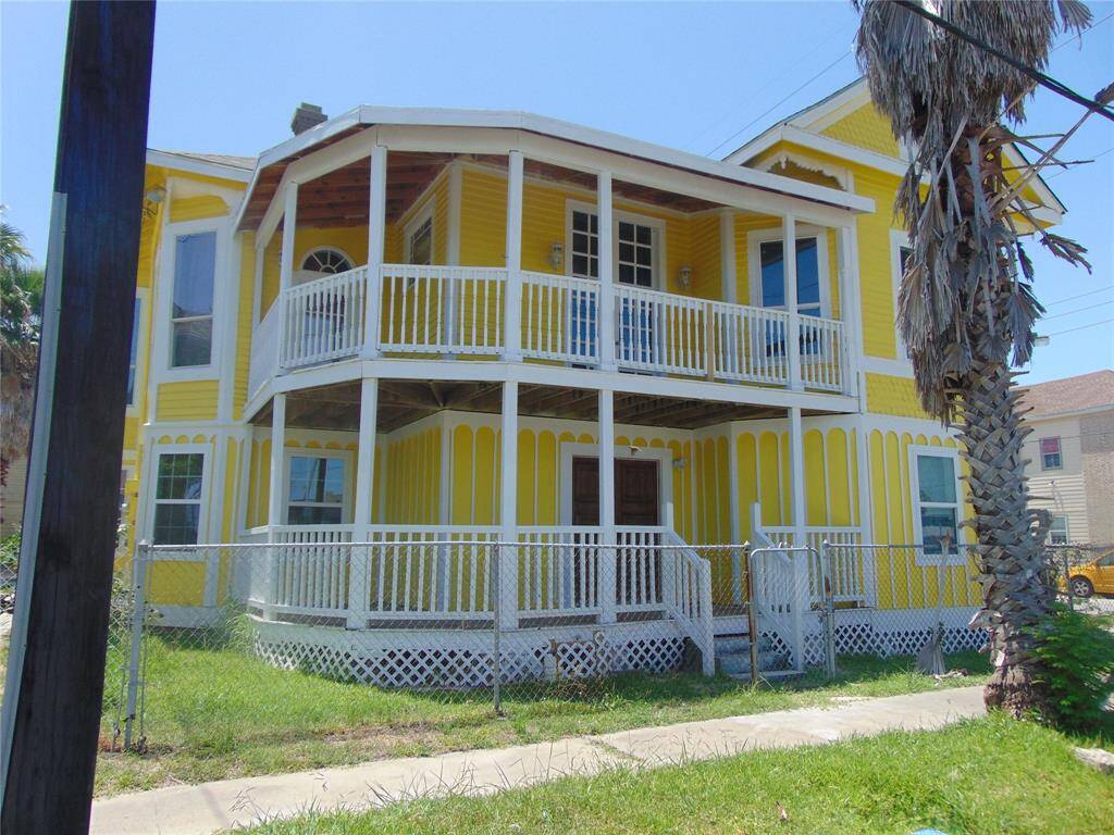 A bright yellow two-story house with charming gingerbread and decorative features on porch of both levels, this property features a small fenced yard accented by palm trees, offering a tropical feel.
