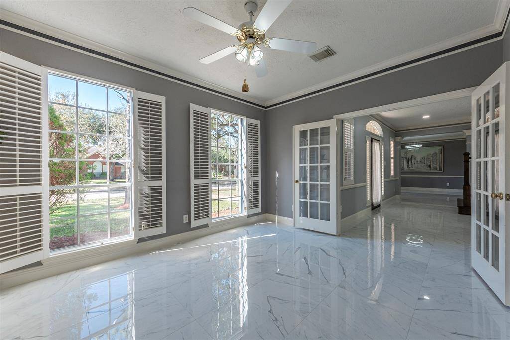 This spacious home-office features custom built-in bookshelf,  elegant porcelain flooring, and large windows with white shutters providing ample natural light.