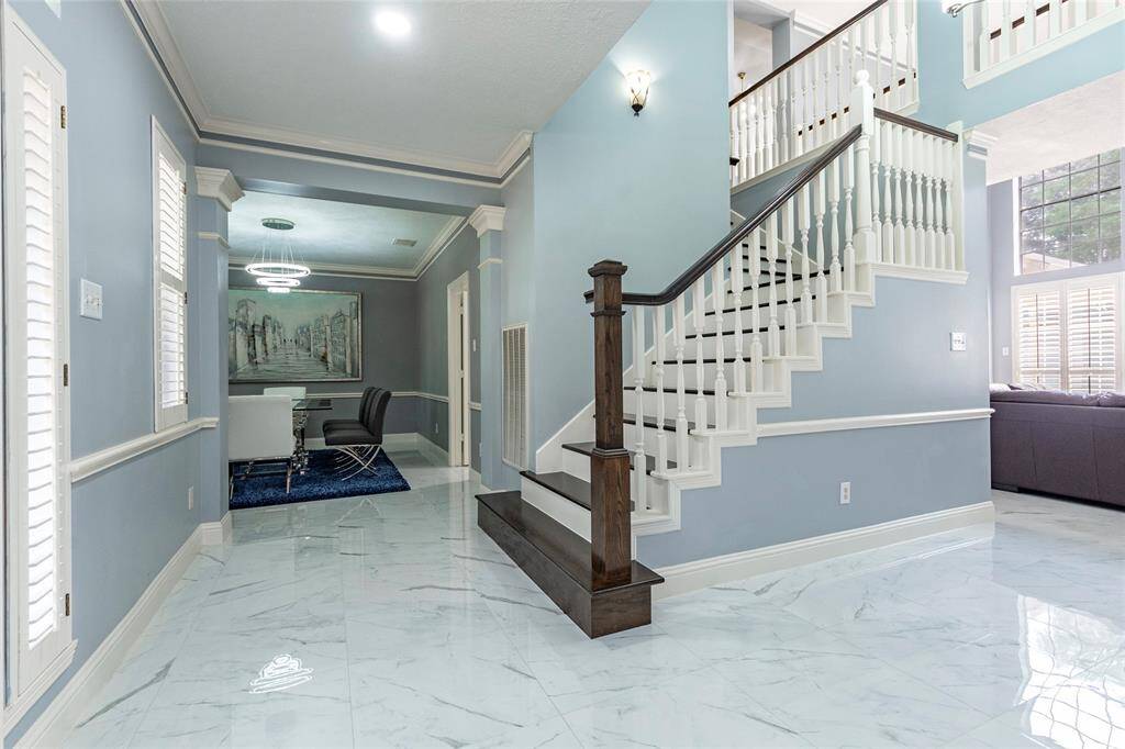 This is a bright and elegant two-story entryway featuring a beautiful staircase with wood accents and white risers, complemented by a stylish wrought-iron door and porcelain flooring, which opens up to a spacious and well-lit interior with a view beyond.
