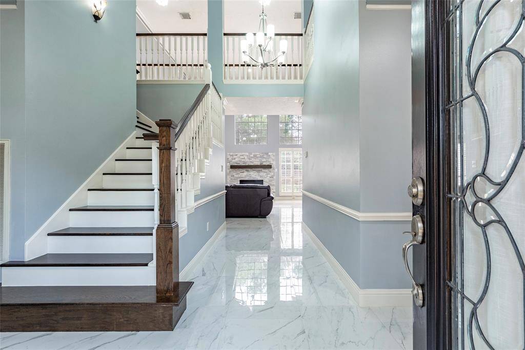 This is an elegant entryway with polished floors, a grand staircase with wood and white balustrades, and soothing neutral paint accented by white trim. The space is well-lit, featuring large windows with plantation shutters and a contemporary chandelier. There is a glimpse into a cozy home office area, suggesting a thoughtful use of space within the home.