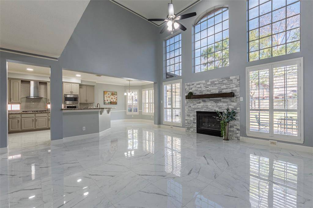 Spacious living room with high ceilings, large windows accented with plantation shutters allowing natural light, a modern stone fireplace, and sleek flooring. I