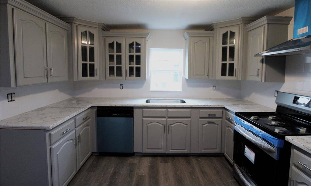 This kitchen features modern gray cabinetry with glass-front upper doors, sleek granite countertops, a stainless steel range, and a dishwasher. It has a central window providing natural light.