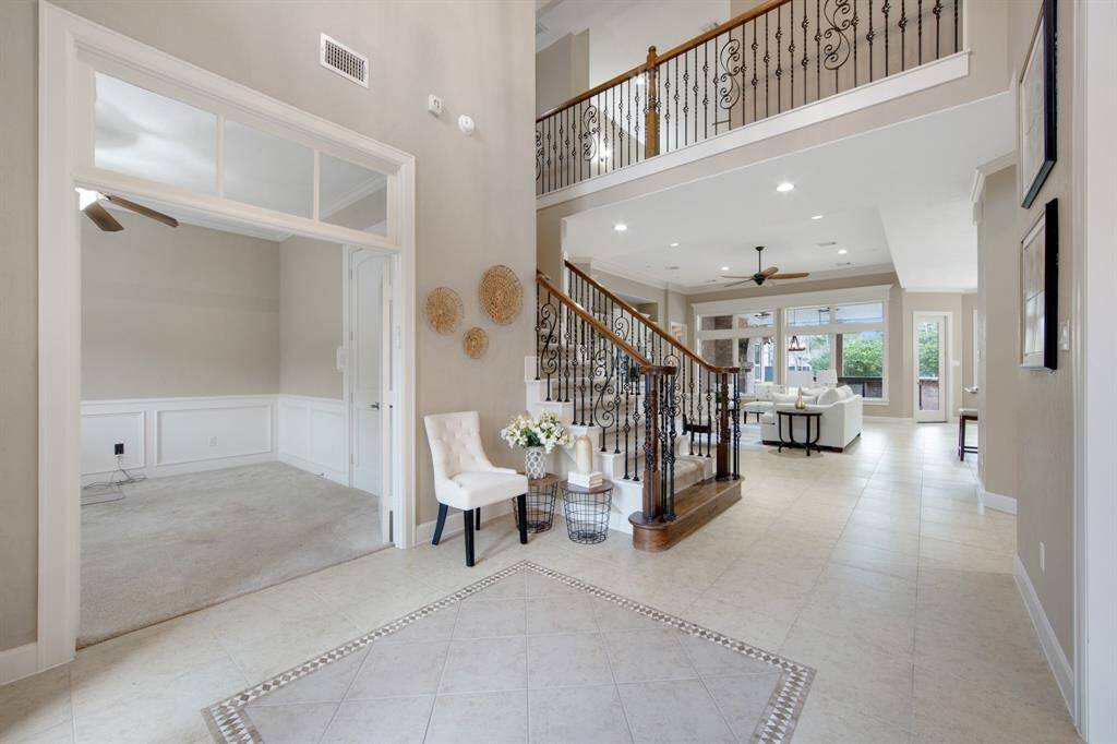 This elegant foyer features high ceilings and a grand staircase with wrought iron railings. The space is filled with natural light from a large window above the front door, and it leads to a bright, open hallway with tile flooring.