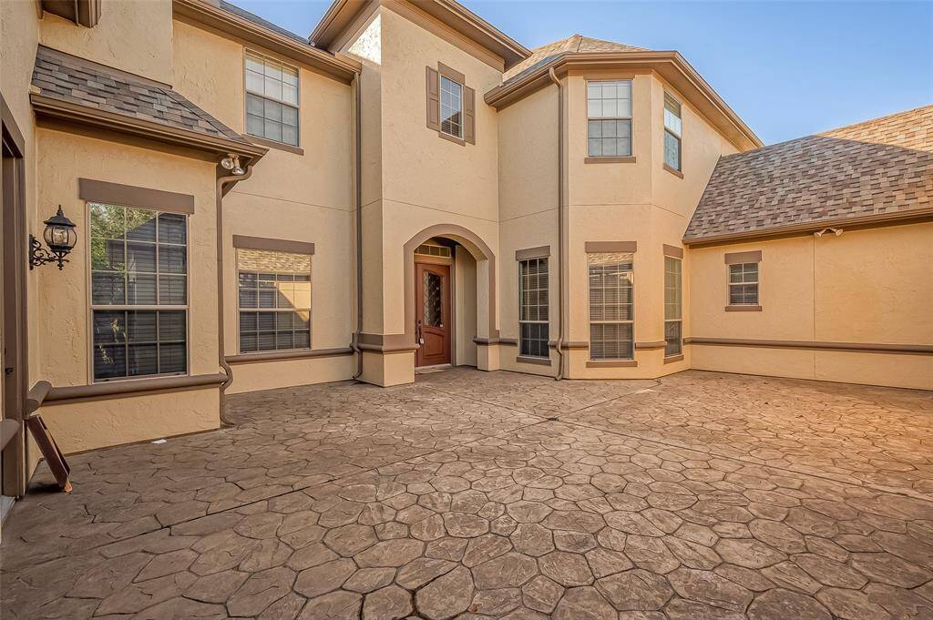 This photo showcases a spacious home exterior with a textured stone courtyard, surrounded by beige stucco walls and multiple large windows. The architecture features a mix of gabled and hip roofing, and there's a prominent wooden front door with an arched entrance, offering a welcoming and elegant appearance.