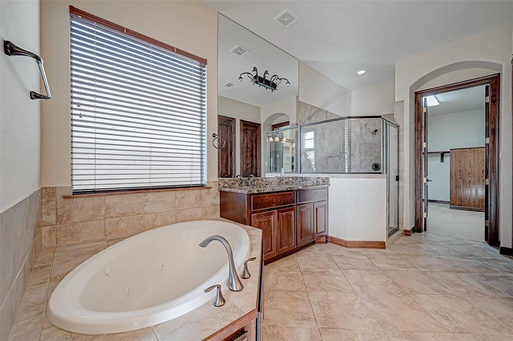 This spacious bathroom features a luxurious soaking tub, a separate glass-enclosed shower, and a granite-topped vanity with ample storage. Natural light streams through a window with blinds, complementing the elegant tile flooring and warm wood accents.