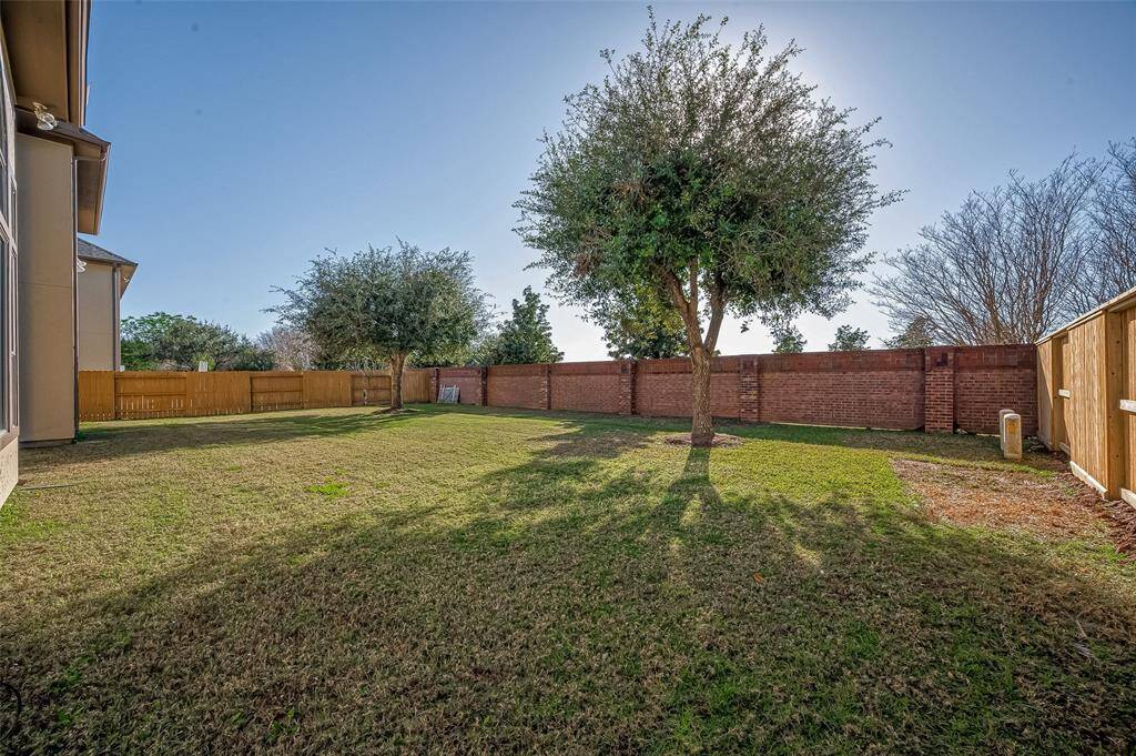 The photo shows a spacious backyard with a well-maintained lawn, bordered by a brick wall and wooden fencing, providing privacy. There are a few mature trees offering shade, creating an inviting outdoor space.