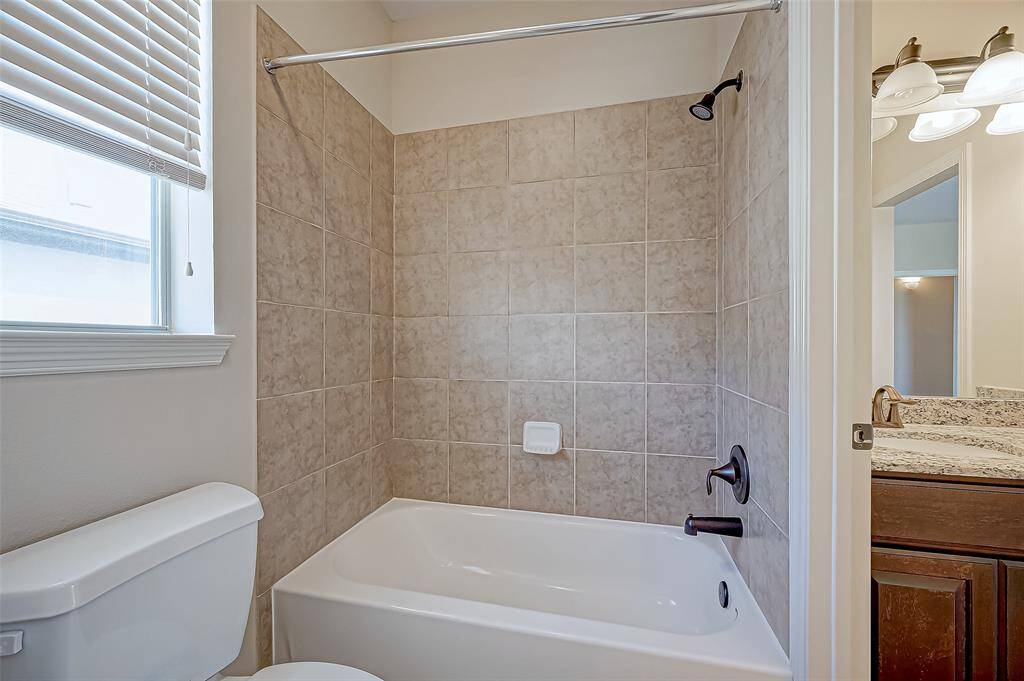 This bathroom features a clean, modern design with a tiled shower and tub combo, a granite countertop, and ample natural light from a window with blinds.