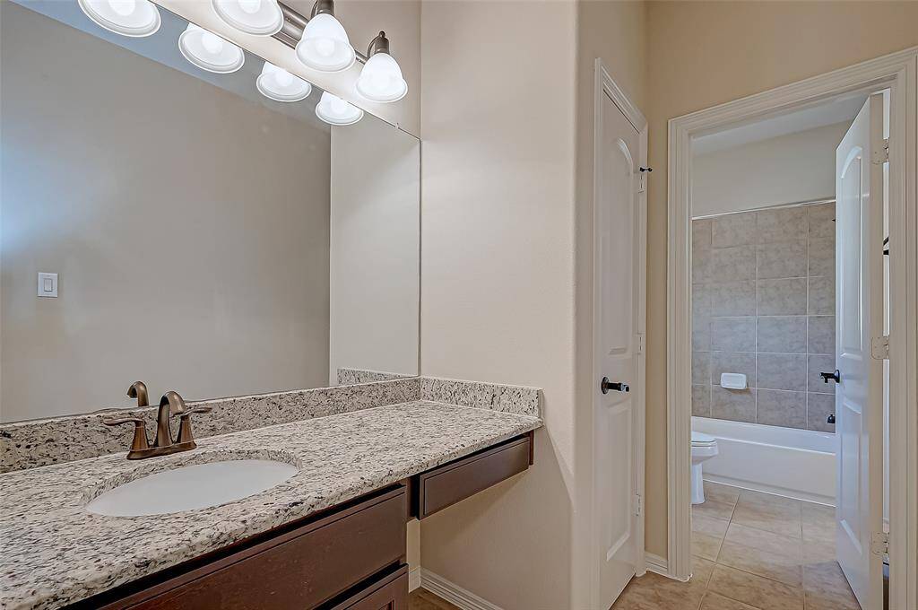 This bathroom features a granite countertop with an undermount sink and elegant fixtures. It has a large mirror with stylish lighting above. The space leads to a tiled shower and bathtub area, offering a clean and modern design.