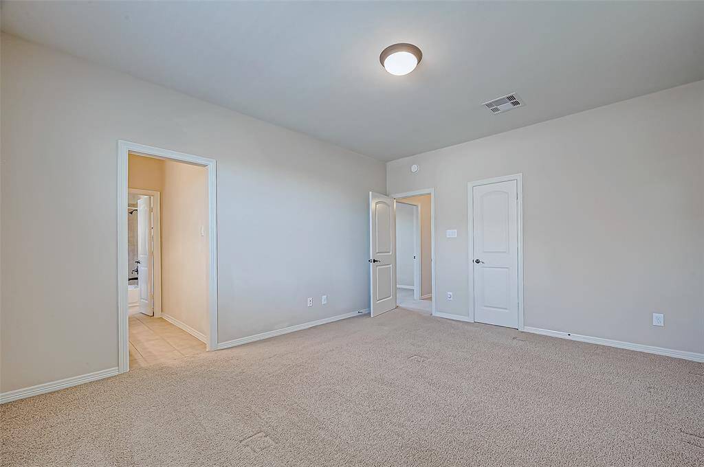 This spacious bedroom features neutral walls, plush carpet, and a ceiling light. It has two doors leading to additional rooms and a bathroom with tile flooring.