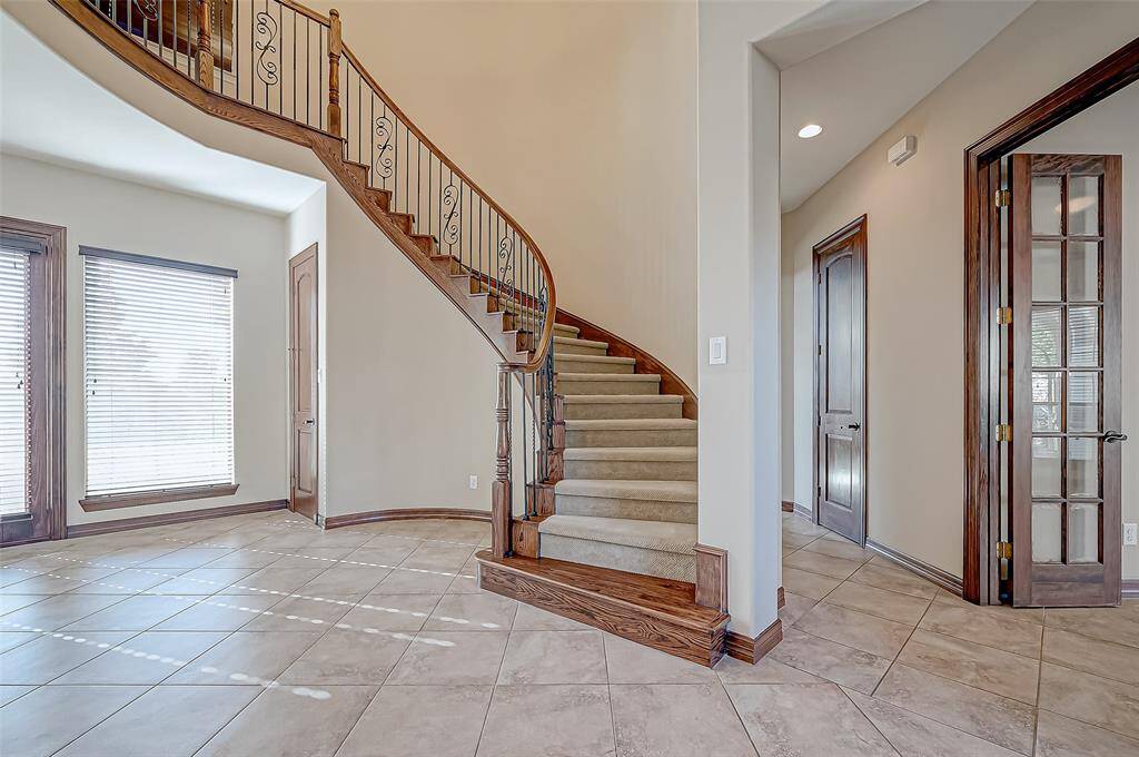 This photo showcases an elegant interior with a grand curved staircase featuring wooden railings and wrought iron balusters. The space is bright with natural light from large windows, tiled floors, and French doors, creating an inviting entryway.