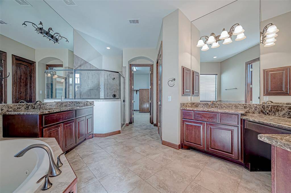 This spacious bathroom features elegant dark wood cabinetry, a large soaking tub, and a glass-enclosed shower. The dual sinks with granite countertops offer ample space, complemented by stylish lighting fixtures and neutral tile flooring.