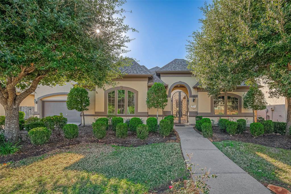 This elegant home features a charming arched entryway with wrought iron detailing, flanked by lush landscaping and mature trees. Large windows provide ample natural light, and the attached garage offers convenience. The well-maintained front yard enhances curb appeal.