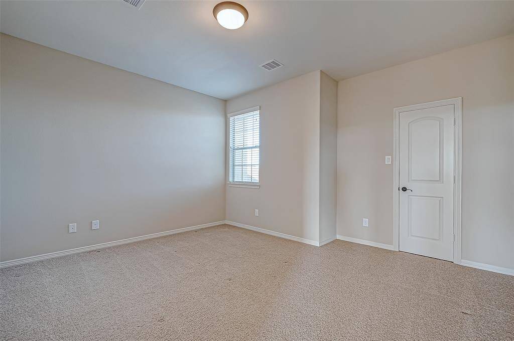 This photo shows a spacious, carpeted room with neutral walls, a single window with blinds, and a ceiling light. The room includes a closed door, offering a clean and versatile space.