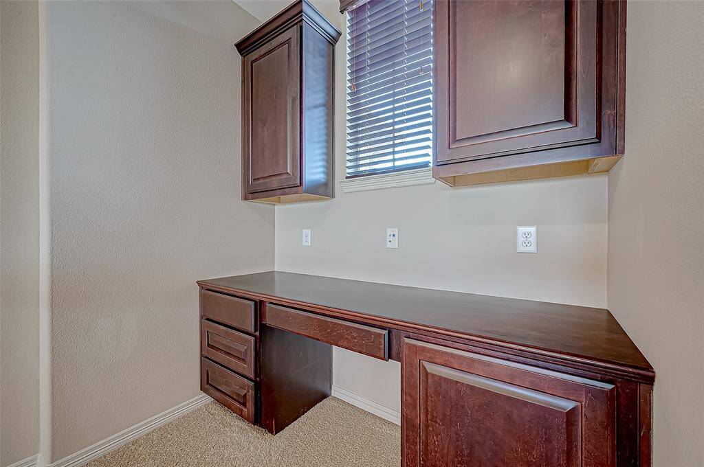 This photo shows a  study corner with dark wood cabinetry and a built-in desk. It features overhead cabinets for storage and a window with blinds for natural light. The setup is ideal for a small workspace or study area.